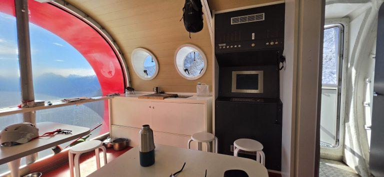 Intérieur du bivouac Gervasutti : coin repas, cuisine et grande baie ouverte sur la vallée