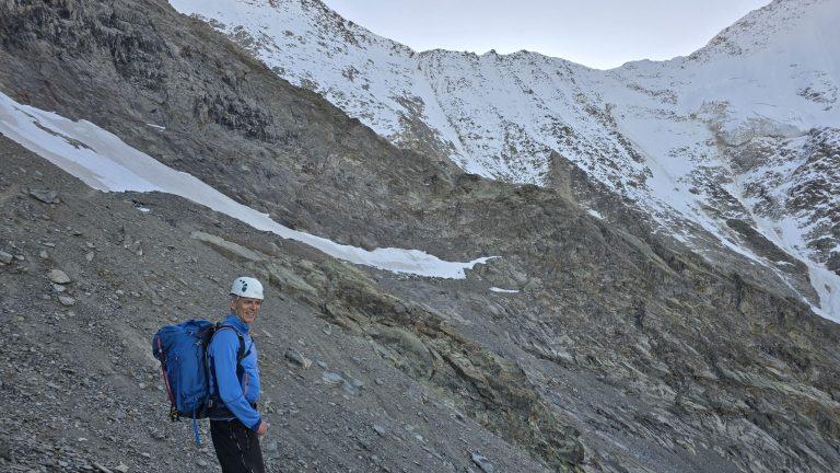 Randonneur casqué au sac bleu dans la pente d'éboulis, sous les faces enneigées