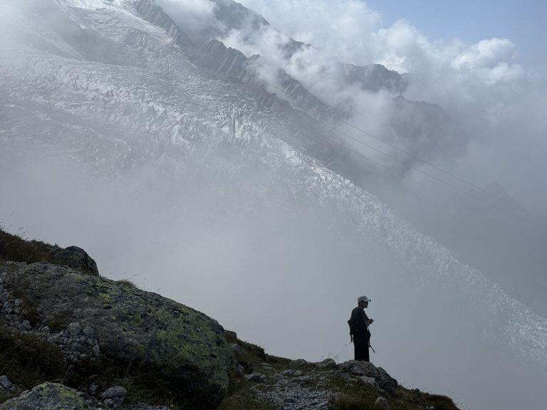 Silhouette d'un randonneur sur un éperon rocheux, face au glacier noyé dans la brume