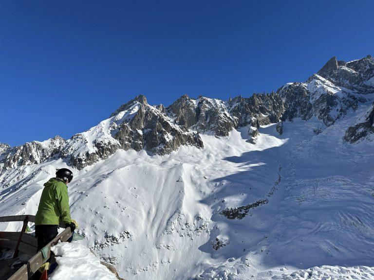 Skieur en veste verte sur la terrasse du refuge, face aux aiguilles enneigées